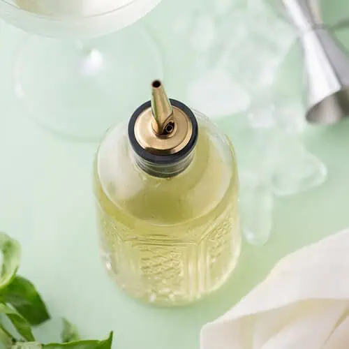 Overhead view of basil simple syrup in a glass bottle with cocktail tools and ice