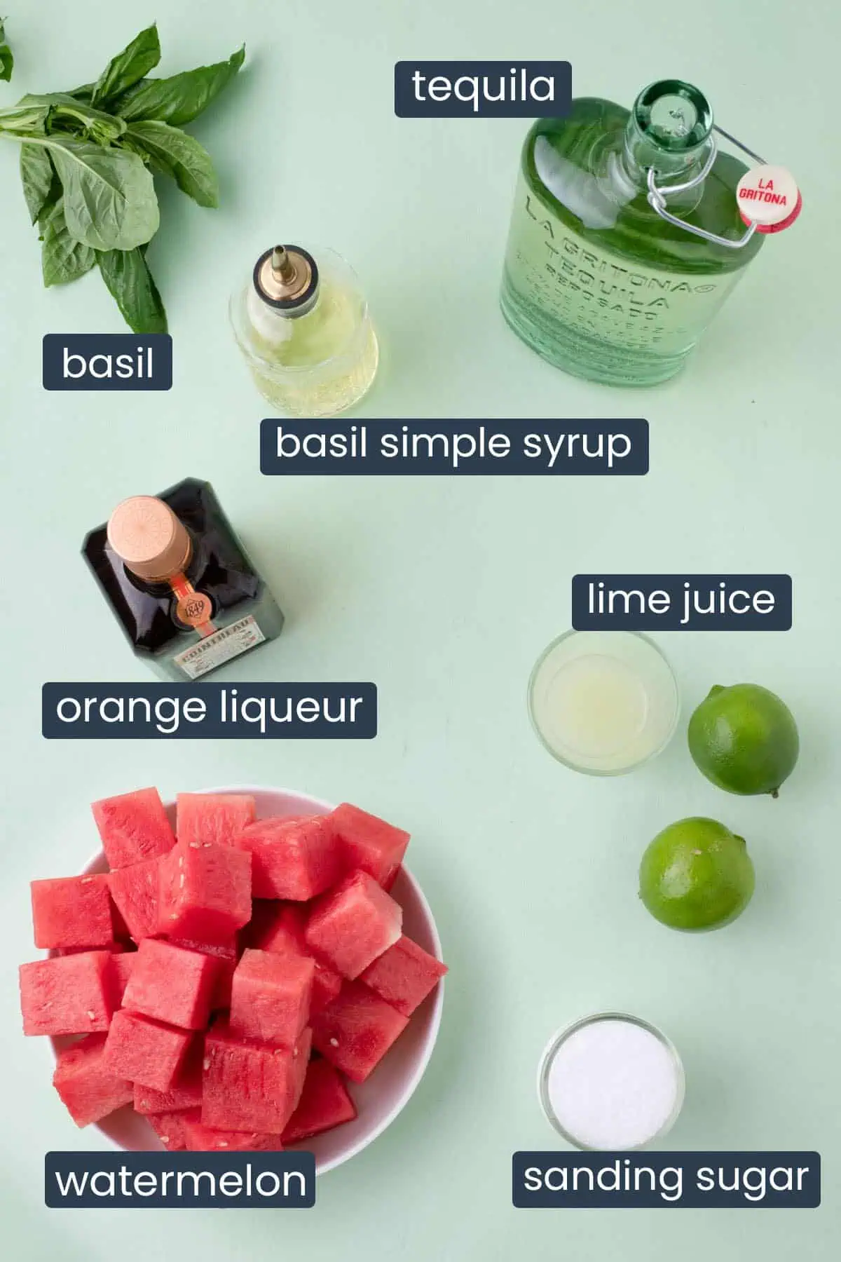 Overhead view of ingredients for a basil watermelon margarita including watermelon, basil, blanco tequila, orange liqueur, lime juice, basil simple syrup, sanding sugar, and limes