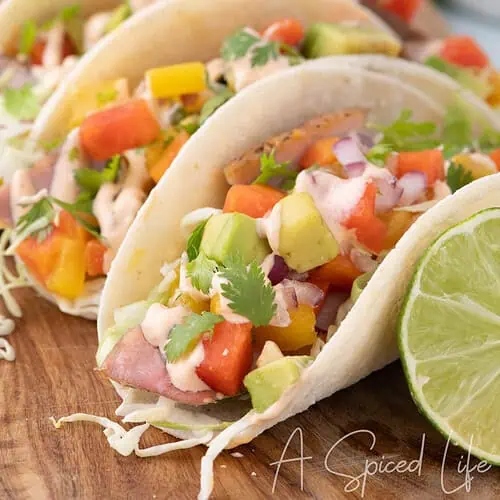 Ahi tuna tacos topped with mango papaya salsa, avocado, cabbage, cilantro, and creamy sriracha sauce beside fresh lime