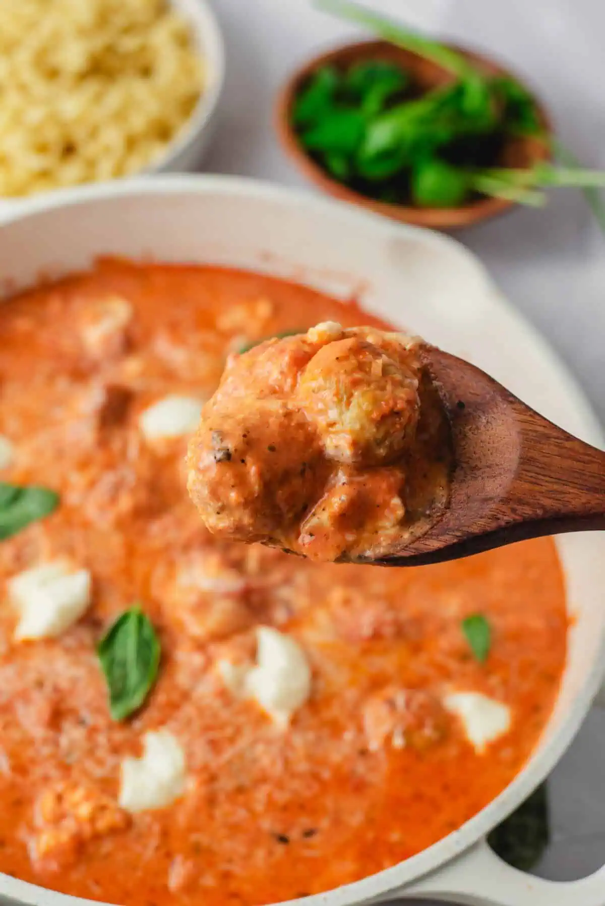 Wooden spoon holding chicken and ricotta meatballs coated in creamy fire-roasted tomato sauce above a skillet of meatballs.