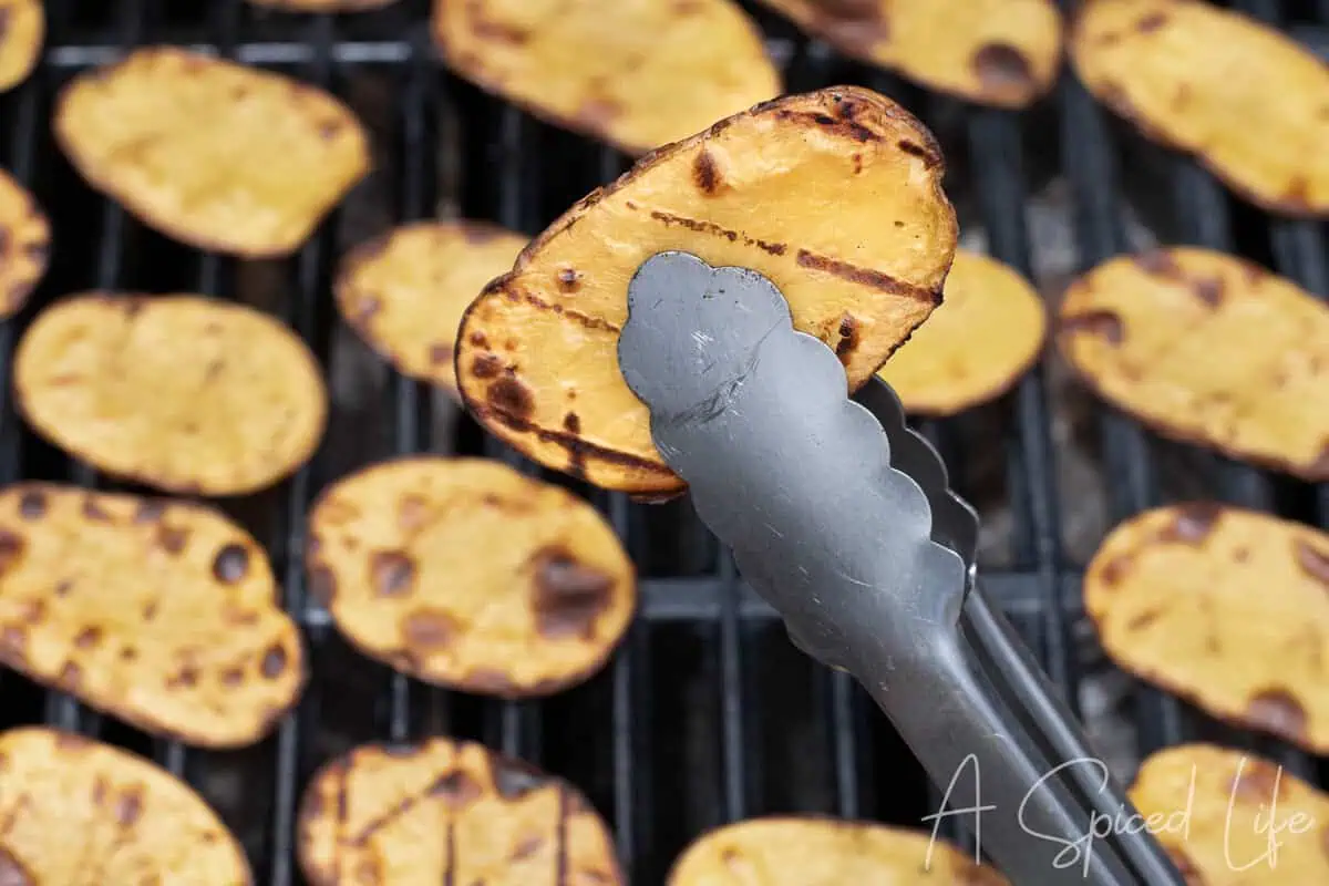 Golden Yukon Gold potato slice held above the grill with metal tongs.