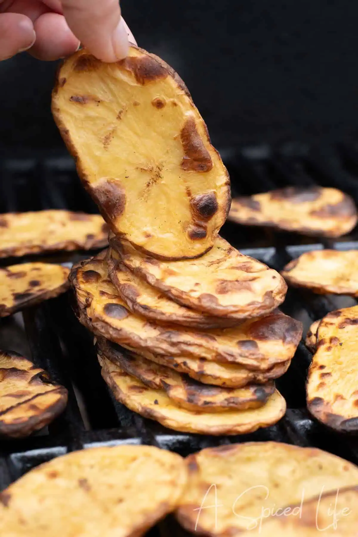A hand lifting a thin, golden potato slice from a stack on the grill.