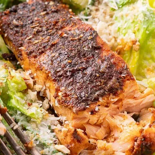Close-up of flaky blackened salmon with a crisp crust served over Caesar salad in a white bowl