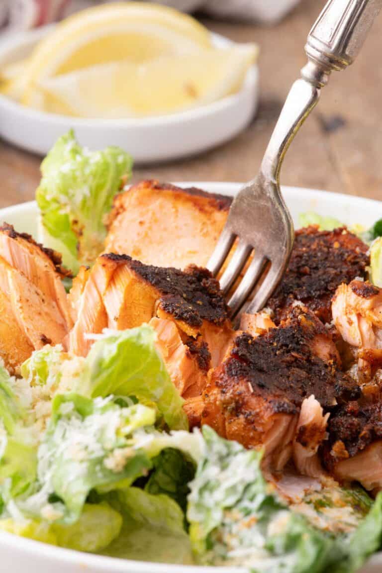 Close-up of flaky blackened salmon served over romaine Caesar salad with Parmesan cheese and creamy dressing.