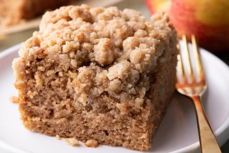 Close-up slice of applesauce crumb cake on a white plate with a thick crumb topping and fresh apples in the background.