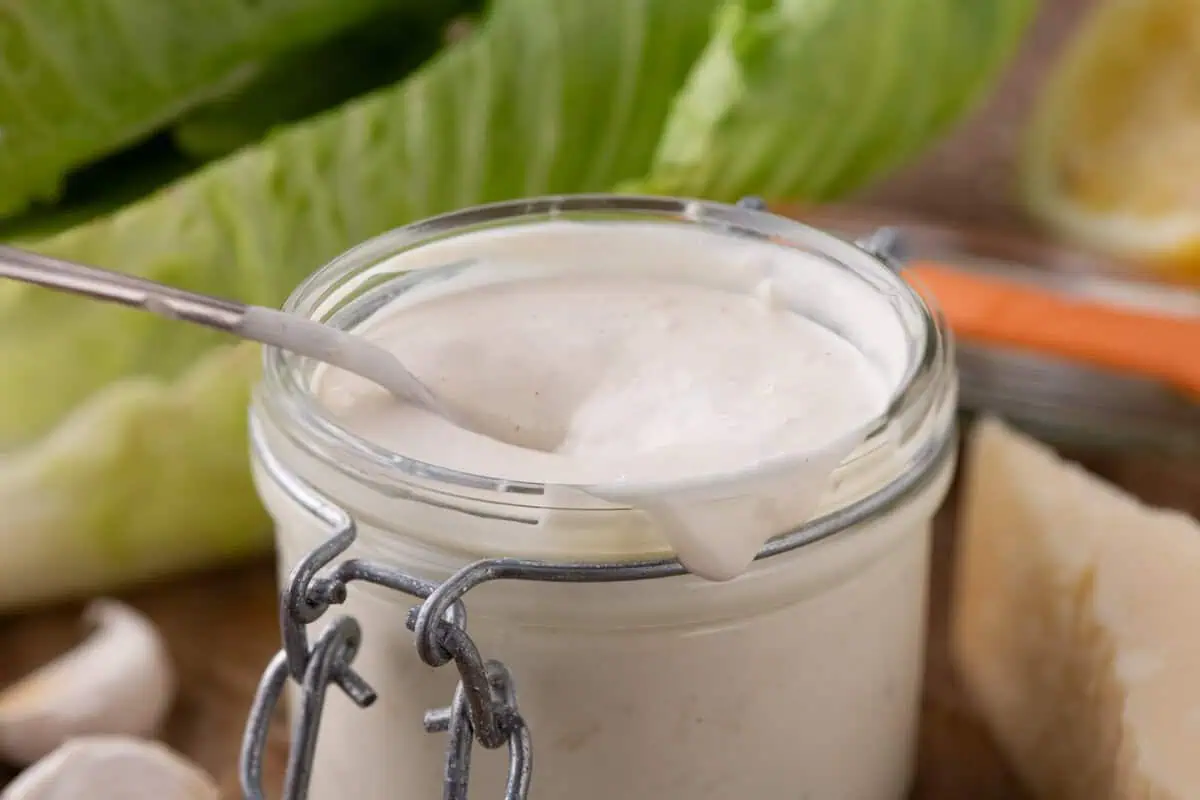 Glass jar filled with creamy Caesar dressing and a spoon, with romaine lettuce, garlic, Parmesan, and lemon nearby.