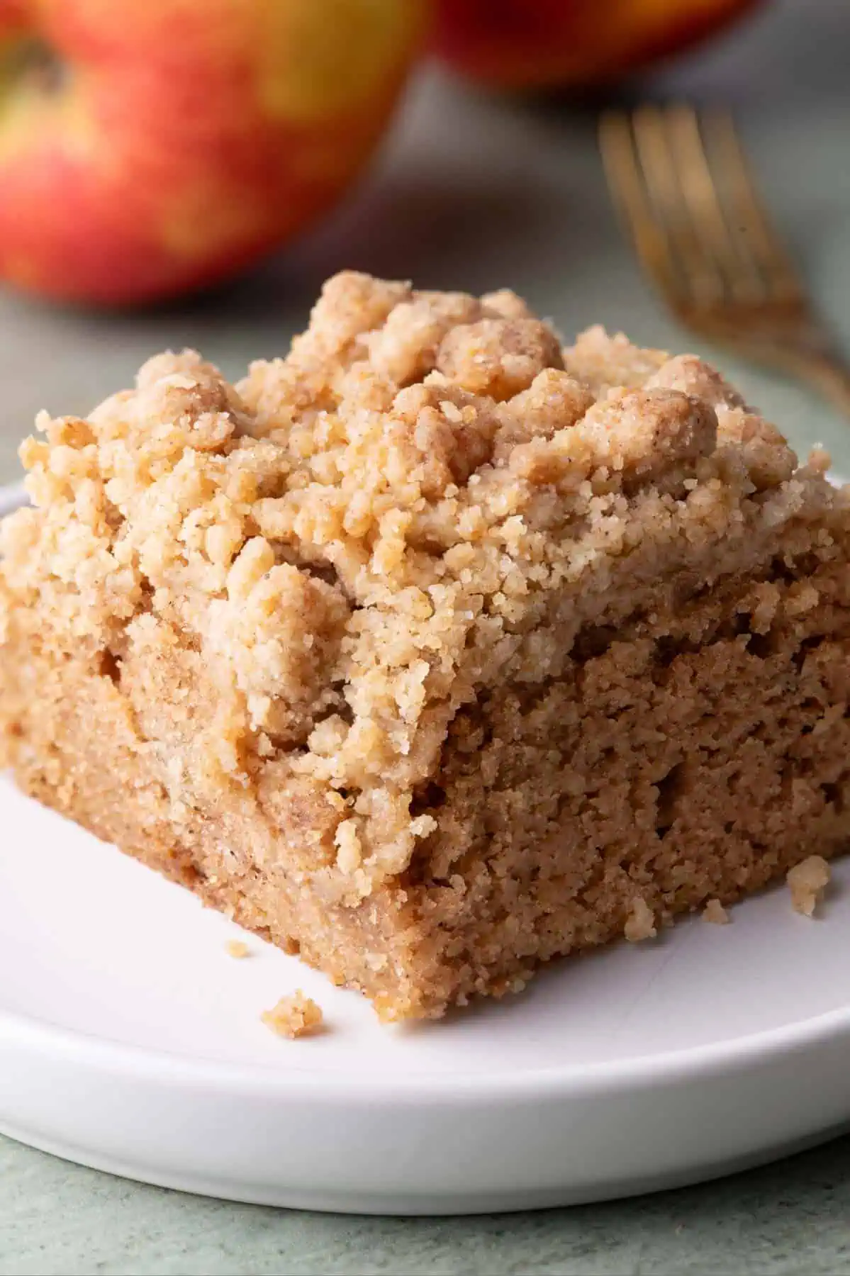Square slice of apple crumb cake with a generous crumb topping on a white plate beside a gold fork and fresh apples.