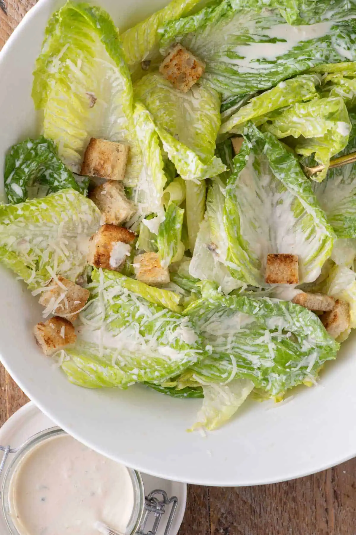 Overhead view of Caesar salad with creamy dressed romaine, croutons, grated Parmesan, and a jar of Caesar dressing at the bottom.