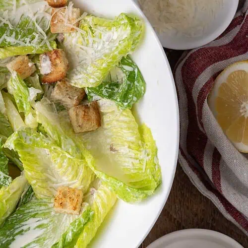 A lighter Caesar salad in a white serving bowl with croutons, Parmesan, and creamy Caesar dressing on the side.