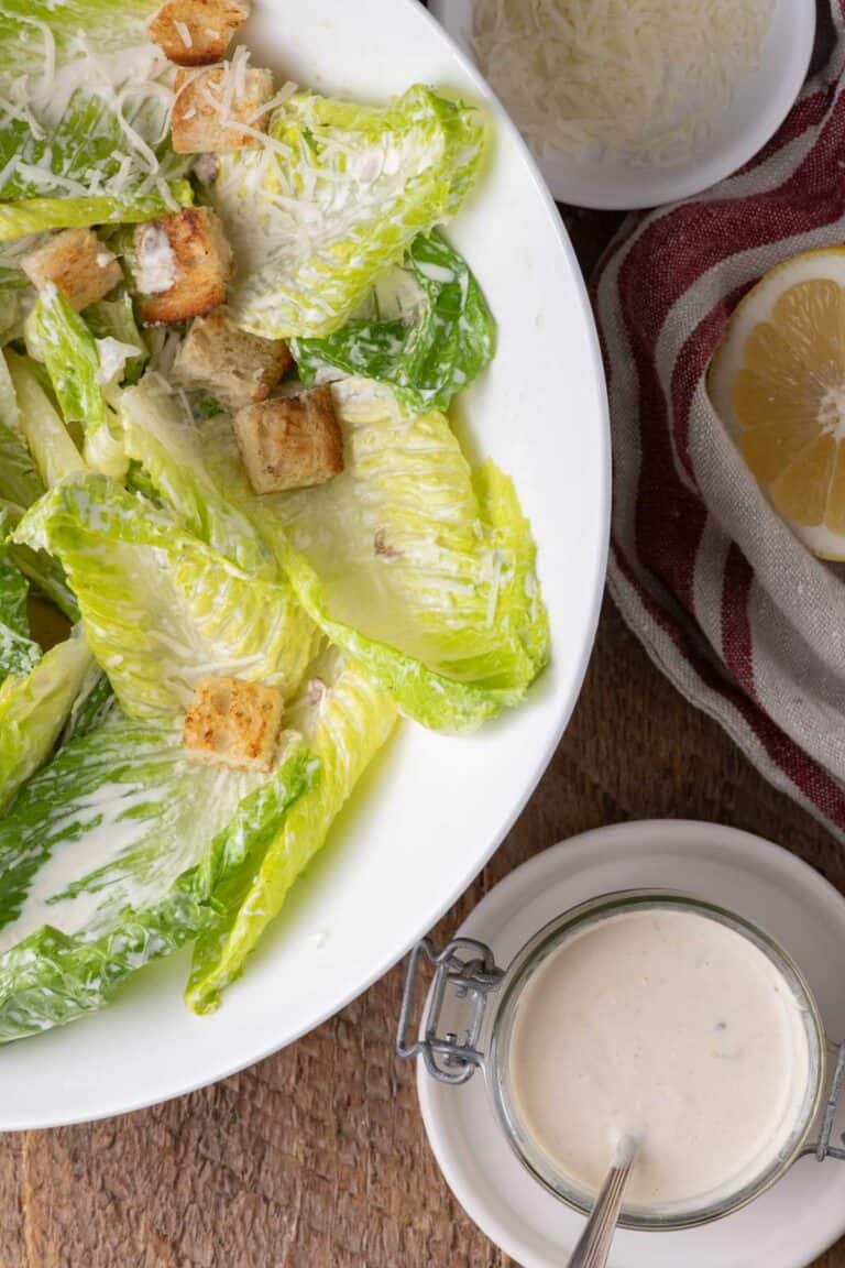 A lighter Caesar salad in a white serving bowl with croutons, Parmesan, and creamy Caesar dressing on the side.
