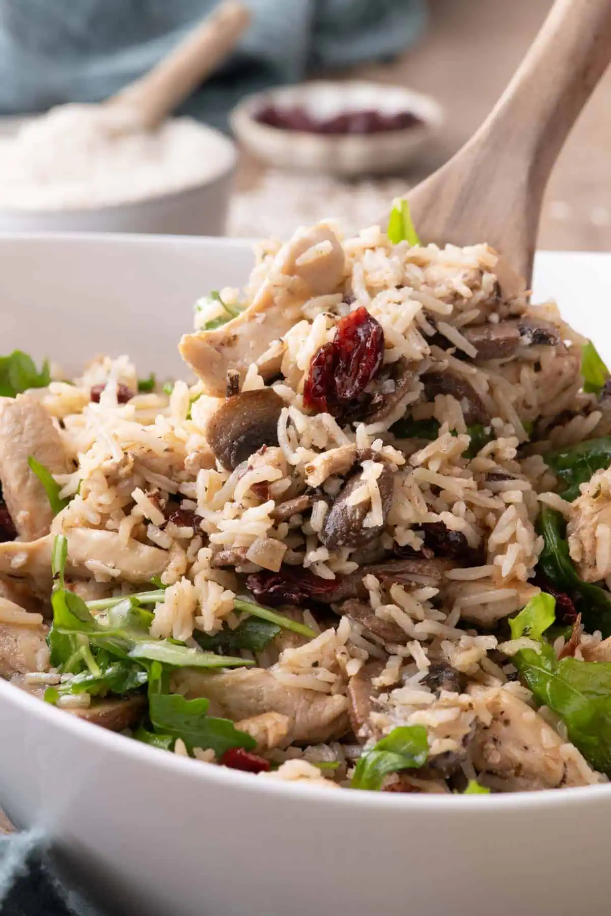Warm chicken and rice salad with mushrooms, cranberries, and arugula in a white bowl with a wooden serving spoon.