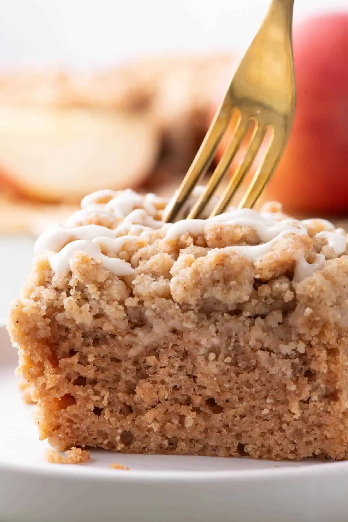 Close-up of glazed apple spice crumb cake with a gold fork pressed into the chunky crumb topping and apples in the background.