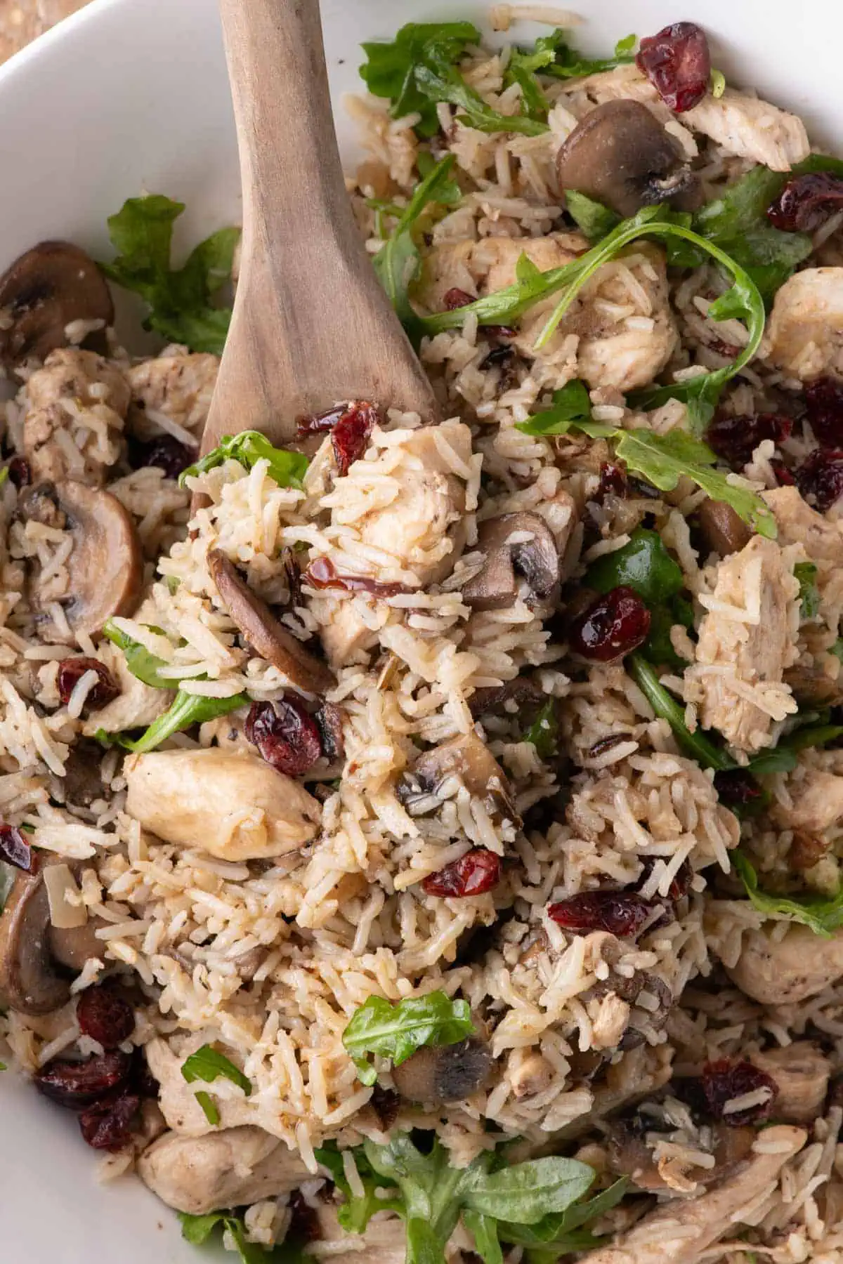 Overhead close-up of chicken and rice salad with mushrooms, cranberries, arugula, and a wooden serving spoon.