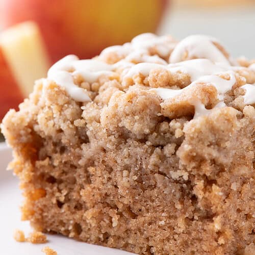 Close-up slice of applesauce crumb cake with chunky cinnamon crumbs and white glaze, with a red apple softly blurred in the background.