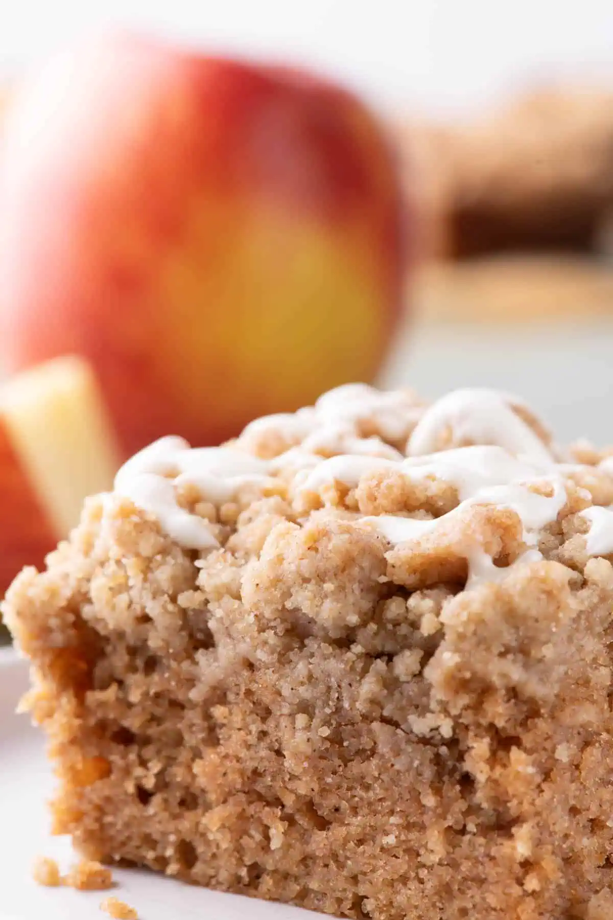 Close-up slice of applesauce crumb cake with chunky cinnamon crumbs and white glaze, with a red apple softly blurred in the background.