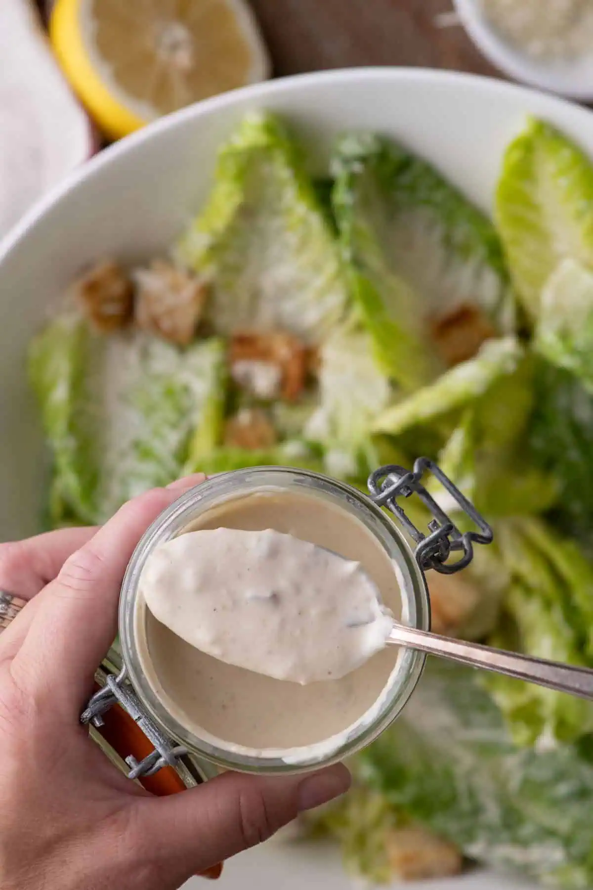 Hand holding a glass jar of creamy Caesar dressing with a spoon, with a bowl of Caesar salad blurred in the background.