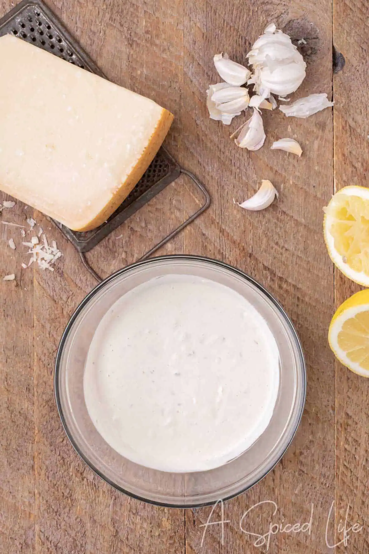 Creamy homemade Caesar dressing mixed in a glass bowl beside Parmesan, garlic and lemon.