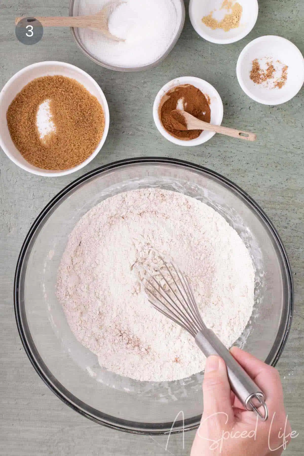 Dry ingredients for the cake batter whisked together in a large glass mixing bowl with small bowls of sugar and spices nearby.