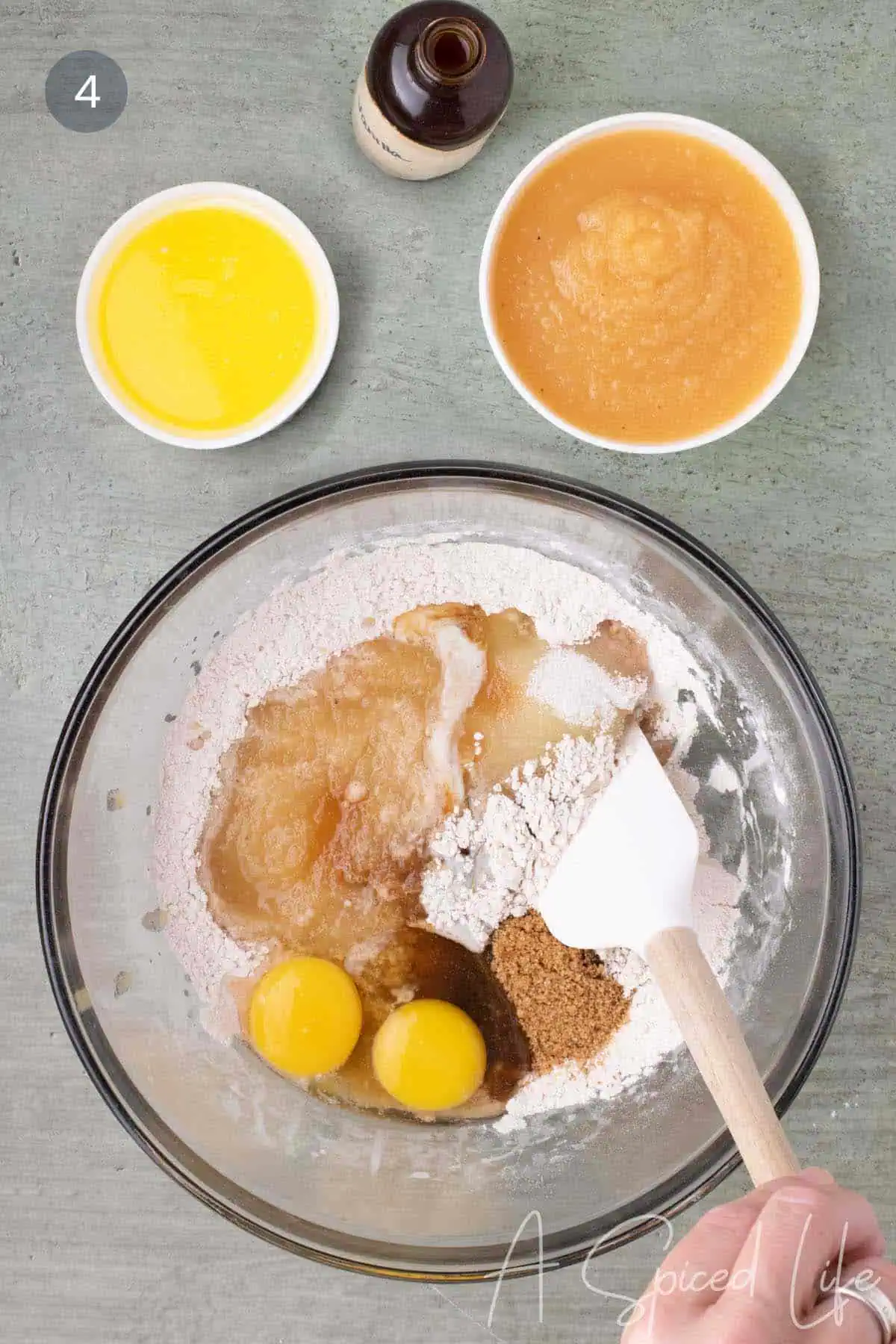 Wet ingredients including applesauce, eggs, vanilla, sugar, and melted butter added to the dry ingredients in a glass mixing bowl.