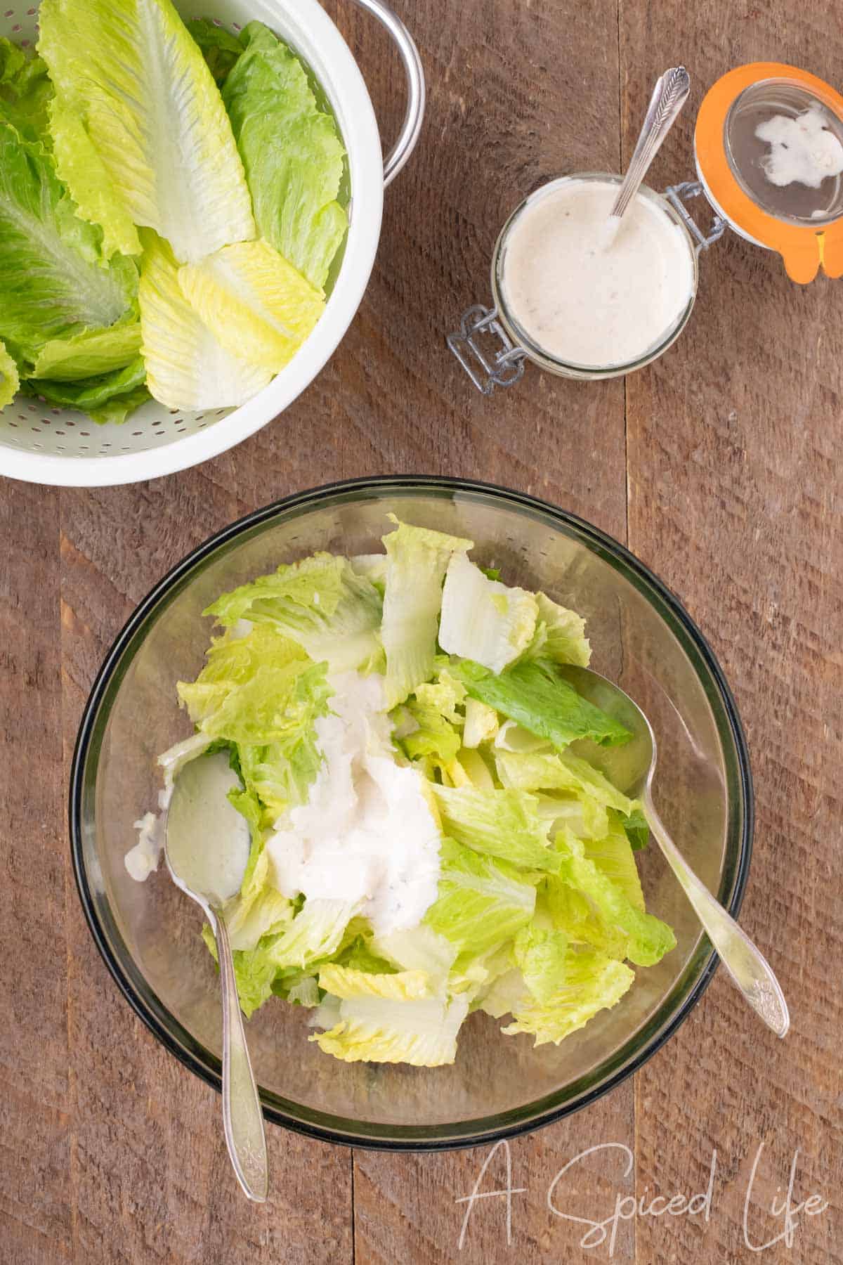 Chopped romaine lettuce being tossed with creamy Caesar dressing in a large glass bowl, with reserved inner leaves nearby.
