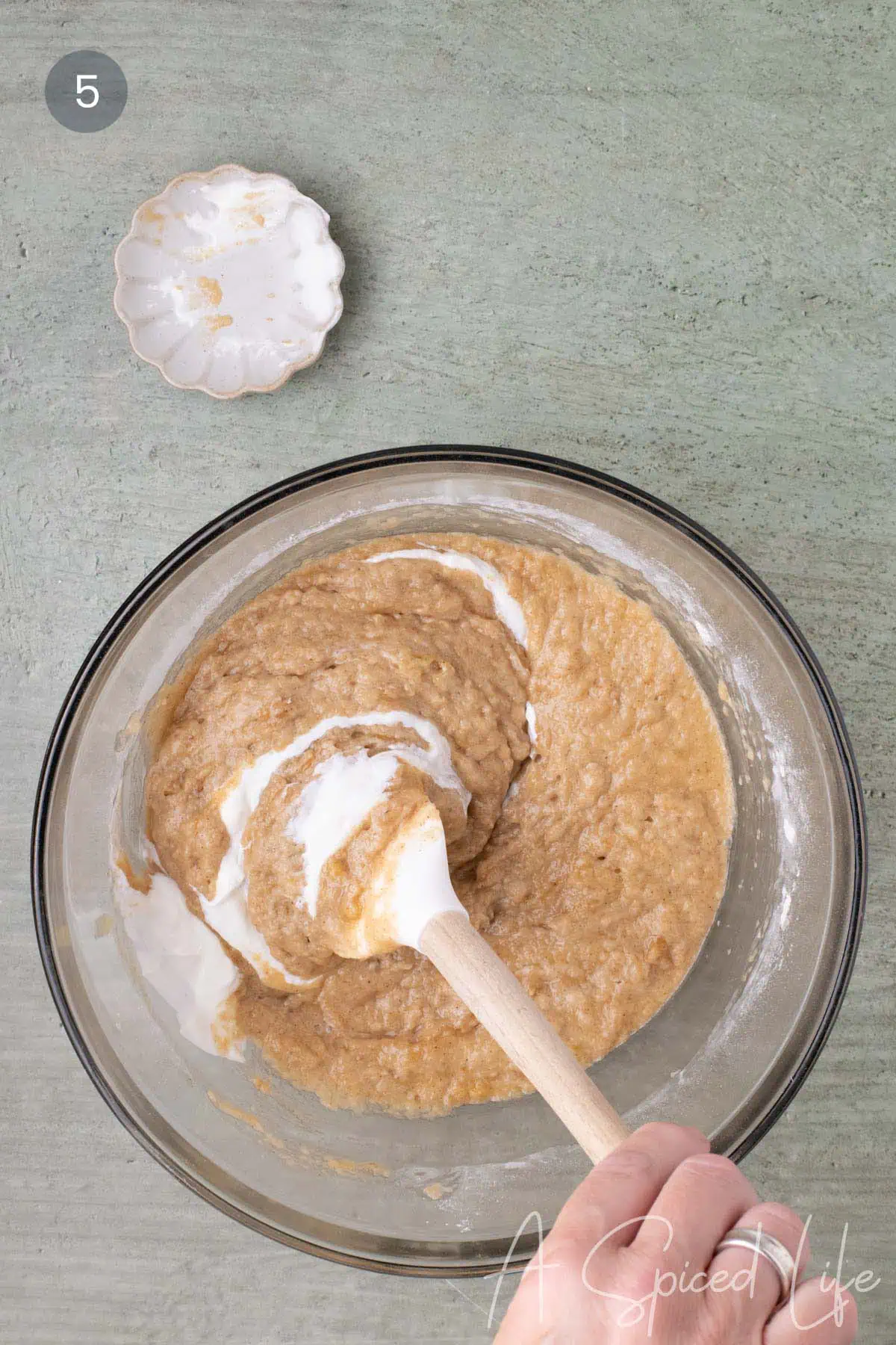 Sour cream being folded into the applesauce cake batter with a spatula in a large glass bowl.