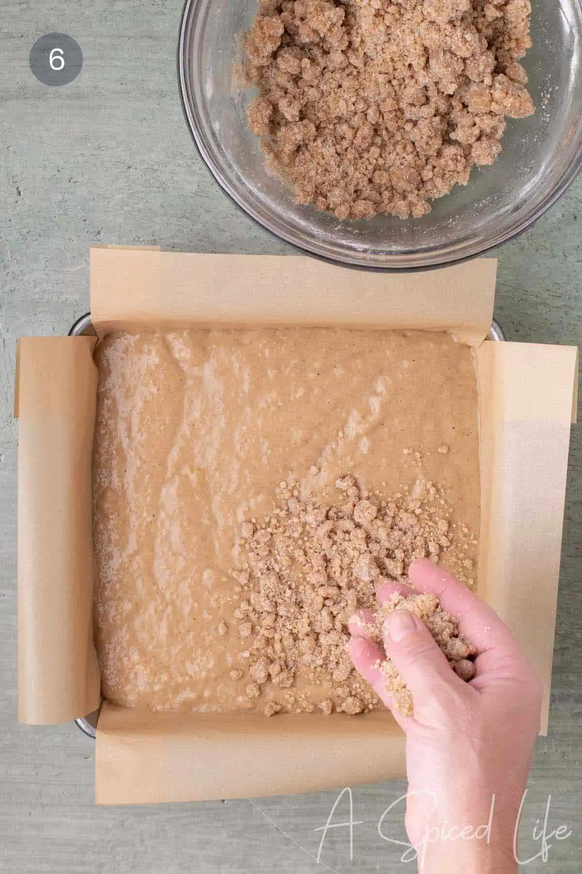 Crumb topping being sprinkled by hand over applesauce cake batter in a parchment-lined 8x8-inch baking pan.