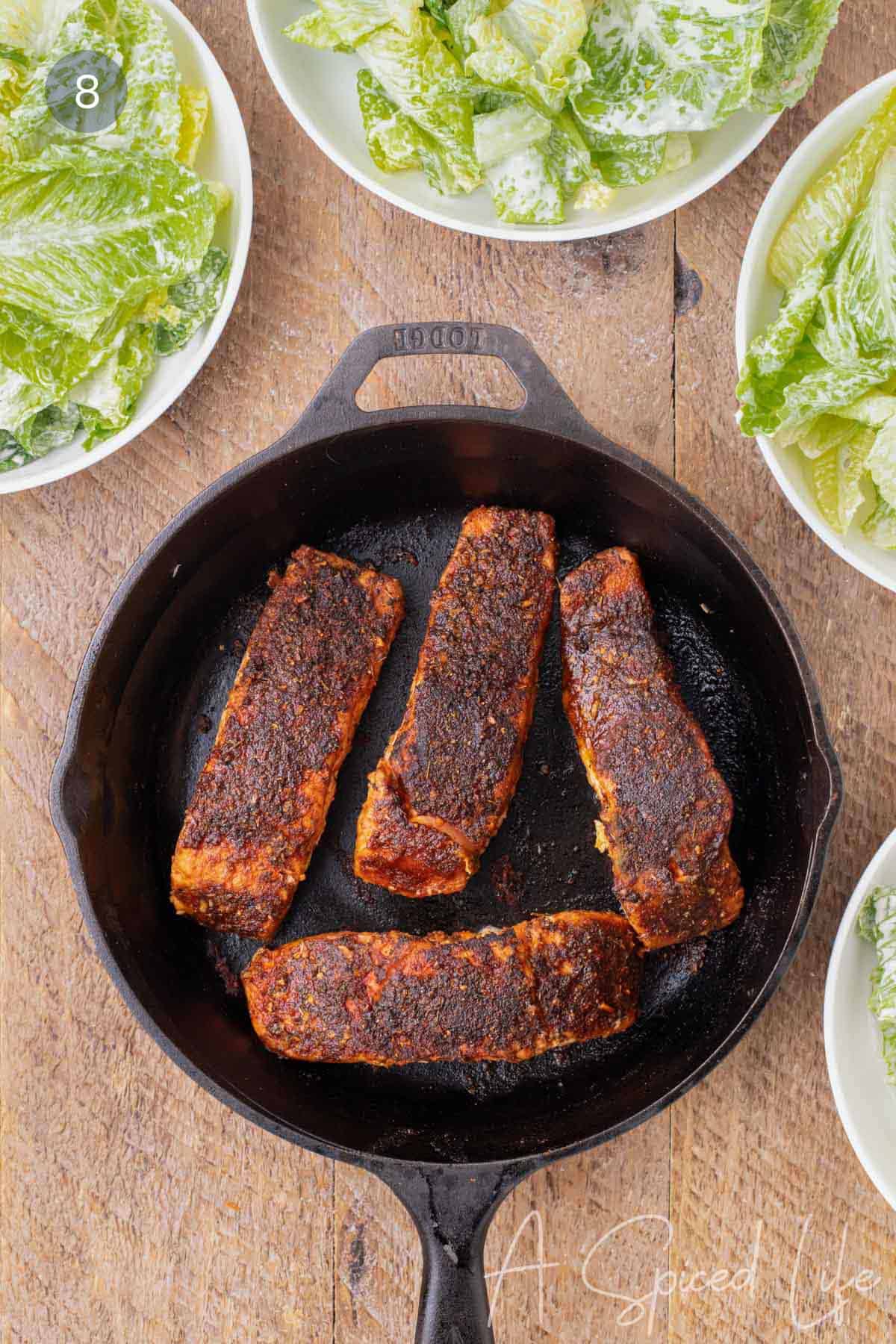 Caesar salad divided among serving bowls while blackened salmon rests nearby.