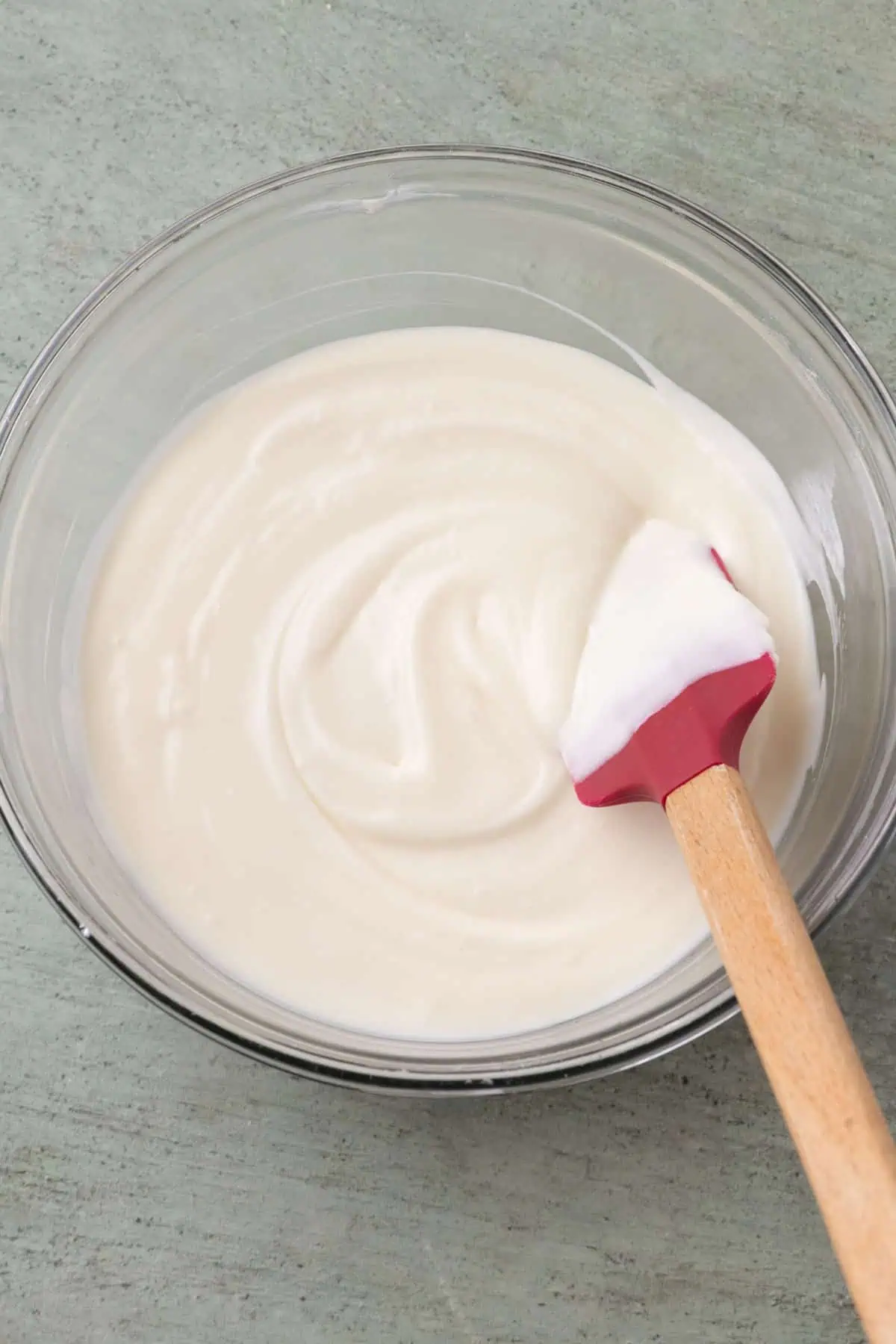 Smooth cream cheese glaze in a glass mixing bowl with a spatula.