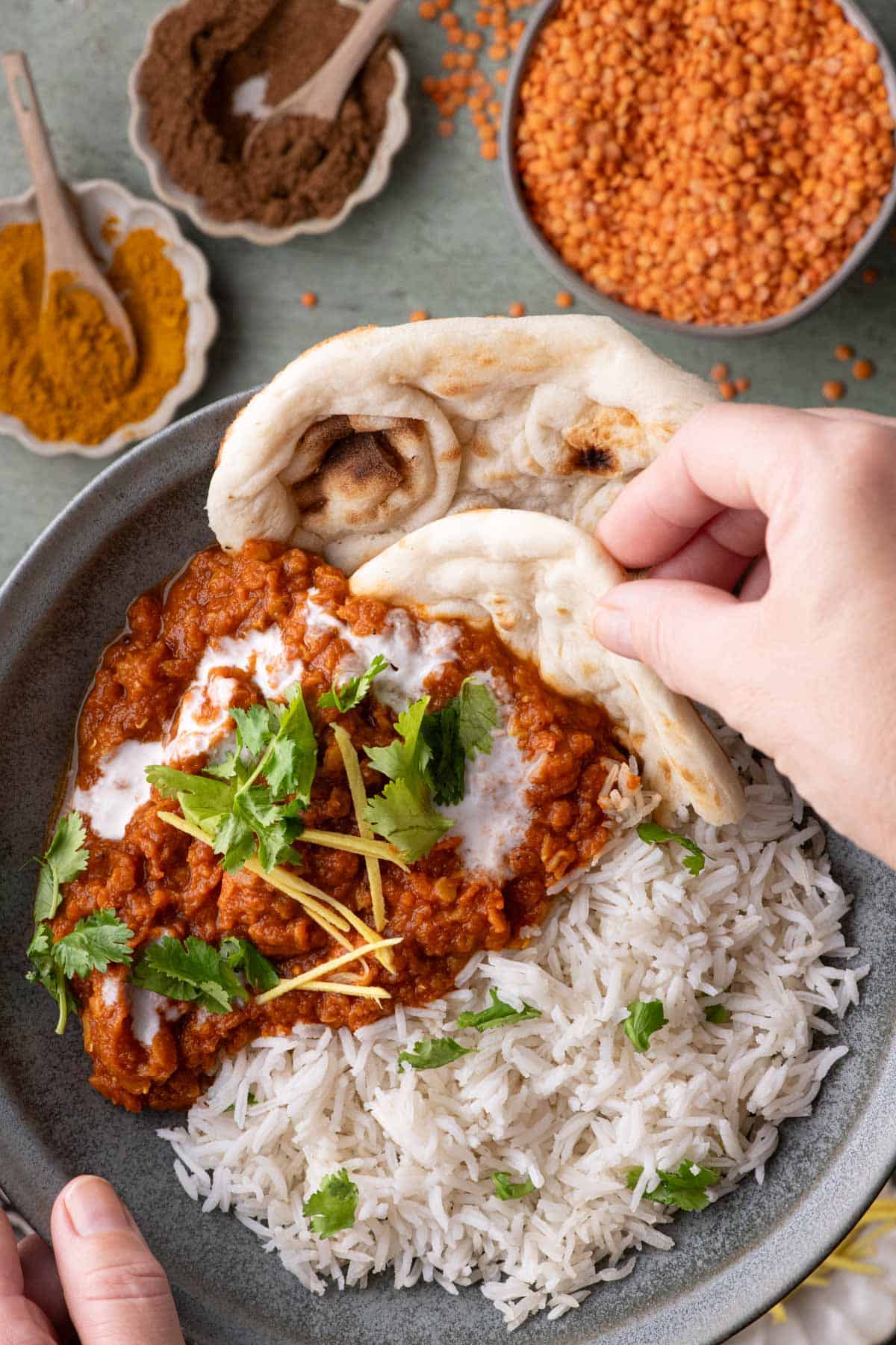 Hand holding naan over a bowl of red lentil stew with rice, coconut cream, cilantro, and fresh ginger