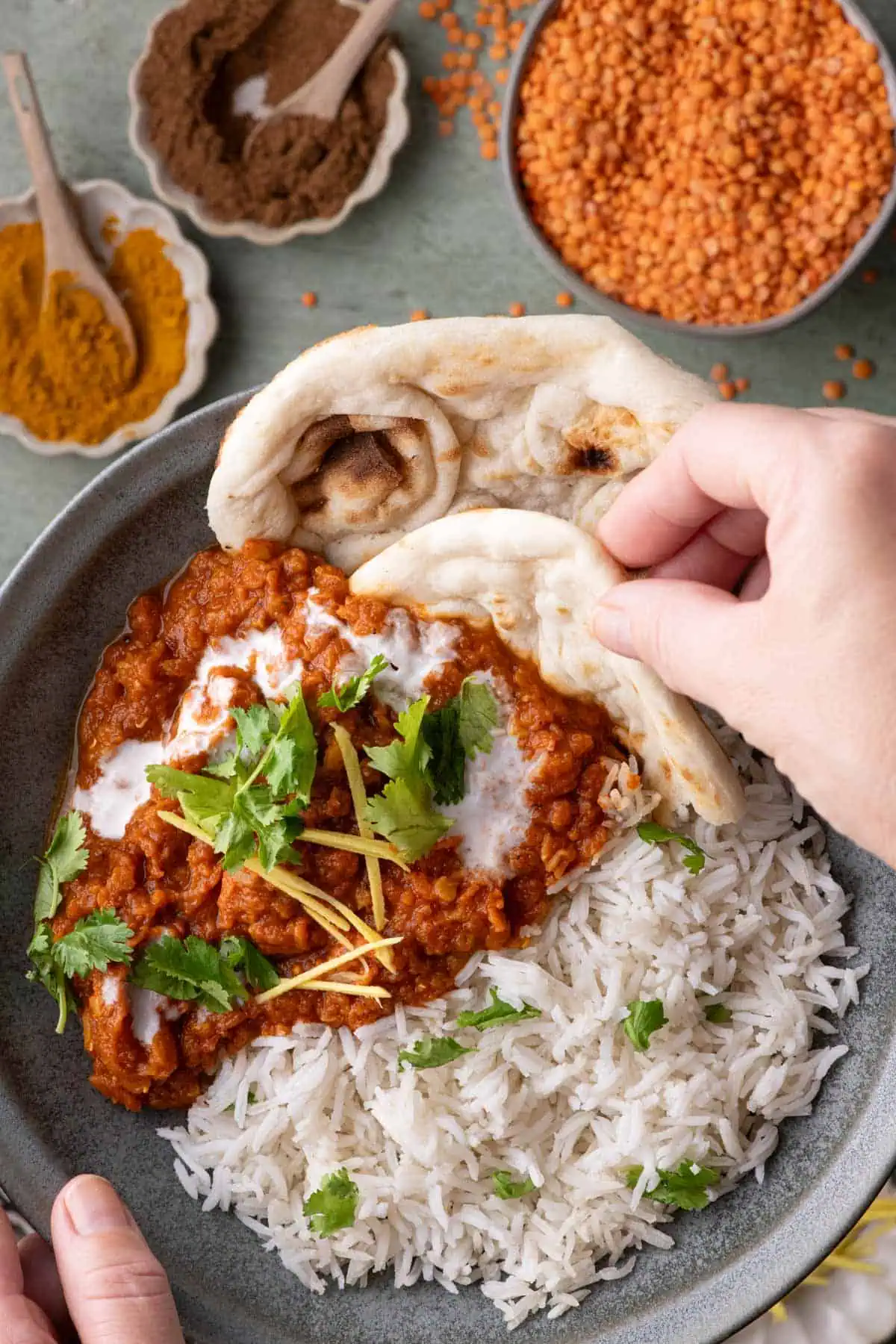 Hand holding naan over a bowl of red lentil stew with rice, coconut cream, cilantro, and fresh ginger