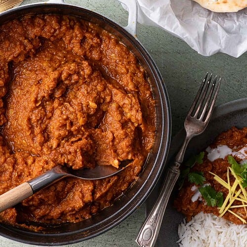 Spiced red lentil stew in a Dutch oven beside a bowl of basmati rice, naan, cilantro, and ginger