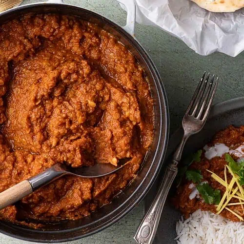 Spiced red lentil stew in a Dutch oven beside a bowl of basmati rice, naan, cilantro, and ginger