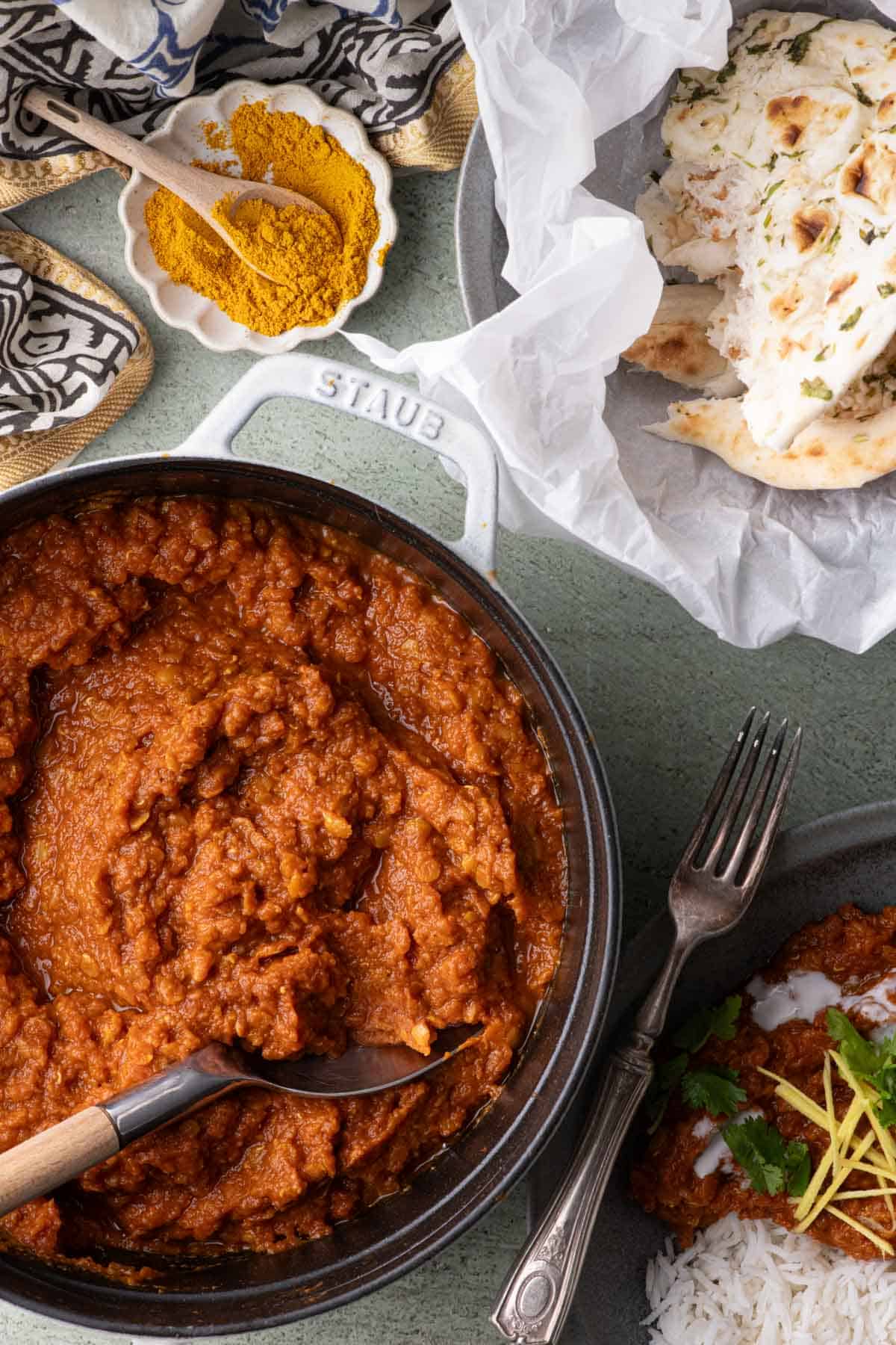 Spiced red lentil stew in a Dutch oven beside a bowl of basmati rice, naan, cilantro, and ginger