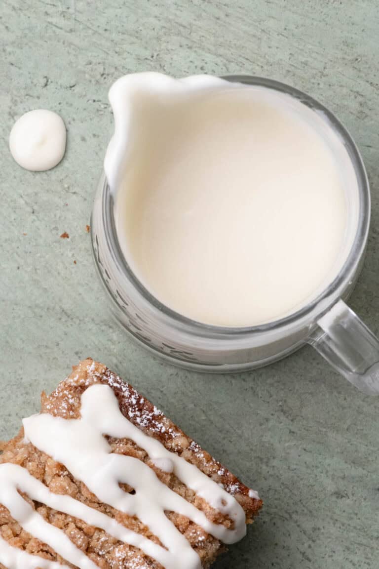 Pitcher of cream cheese glaze beside a slice of glazed crumb cake.