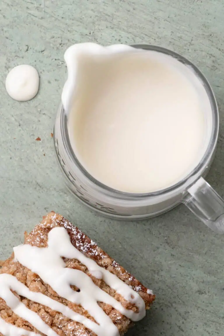 Pitcher of cream cheese glaze beside a slice of glazed crumb cake.