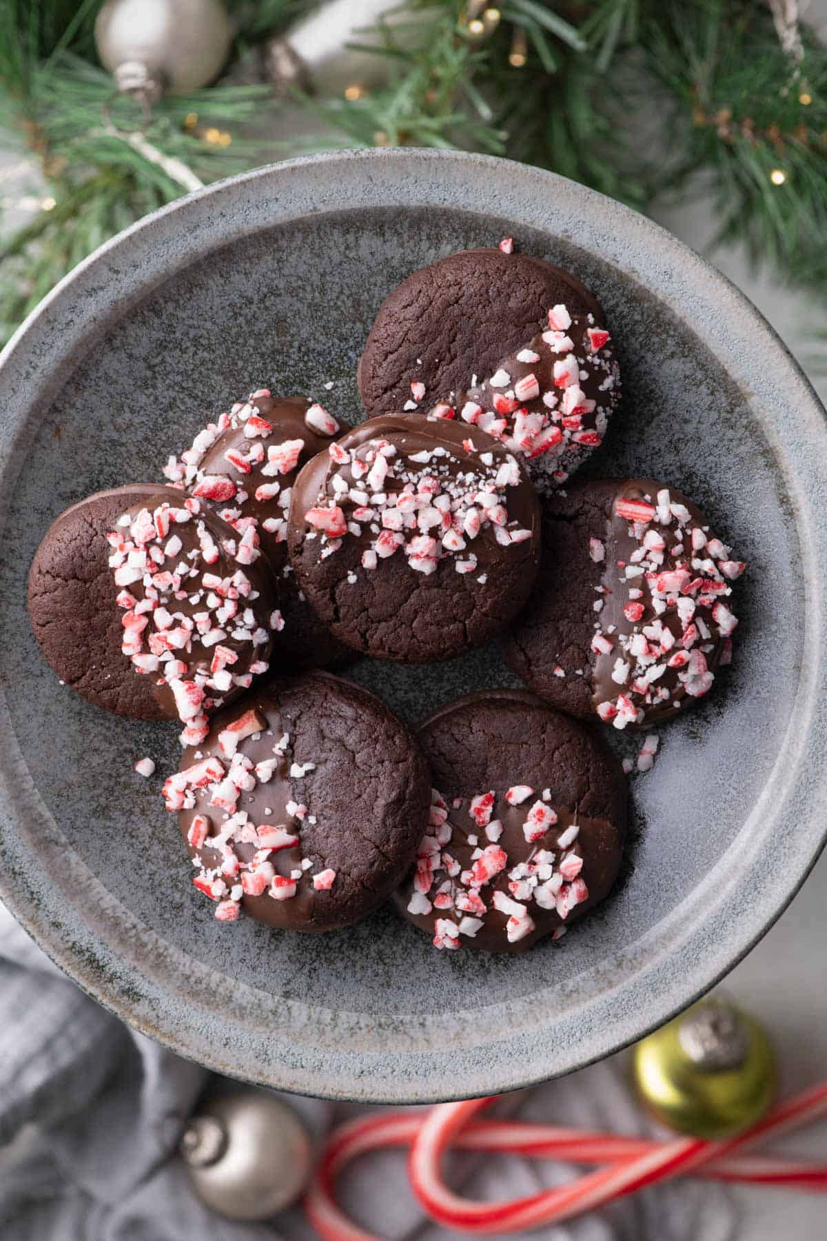 Overhead view of chocolate dipped peppermint cookies arranged on a dark platter with Christmas greenery, ornaments, and candy canes.
