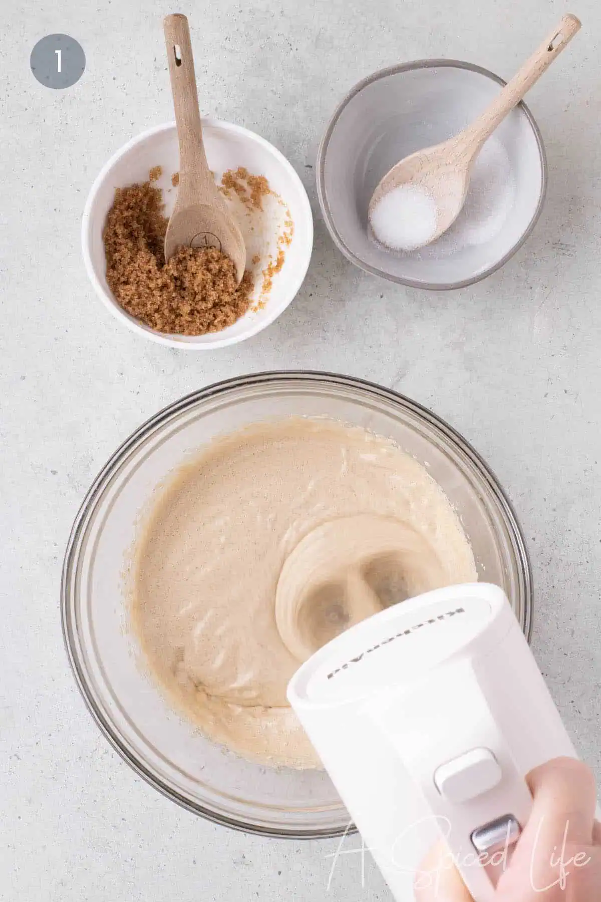 Hand mixer beating an egg and sugar mixture in a glass bowl, with brown sugar and granulated sugar in bowls above