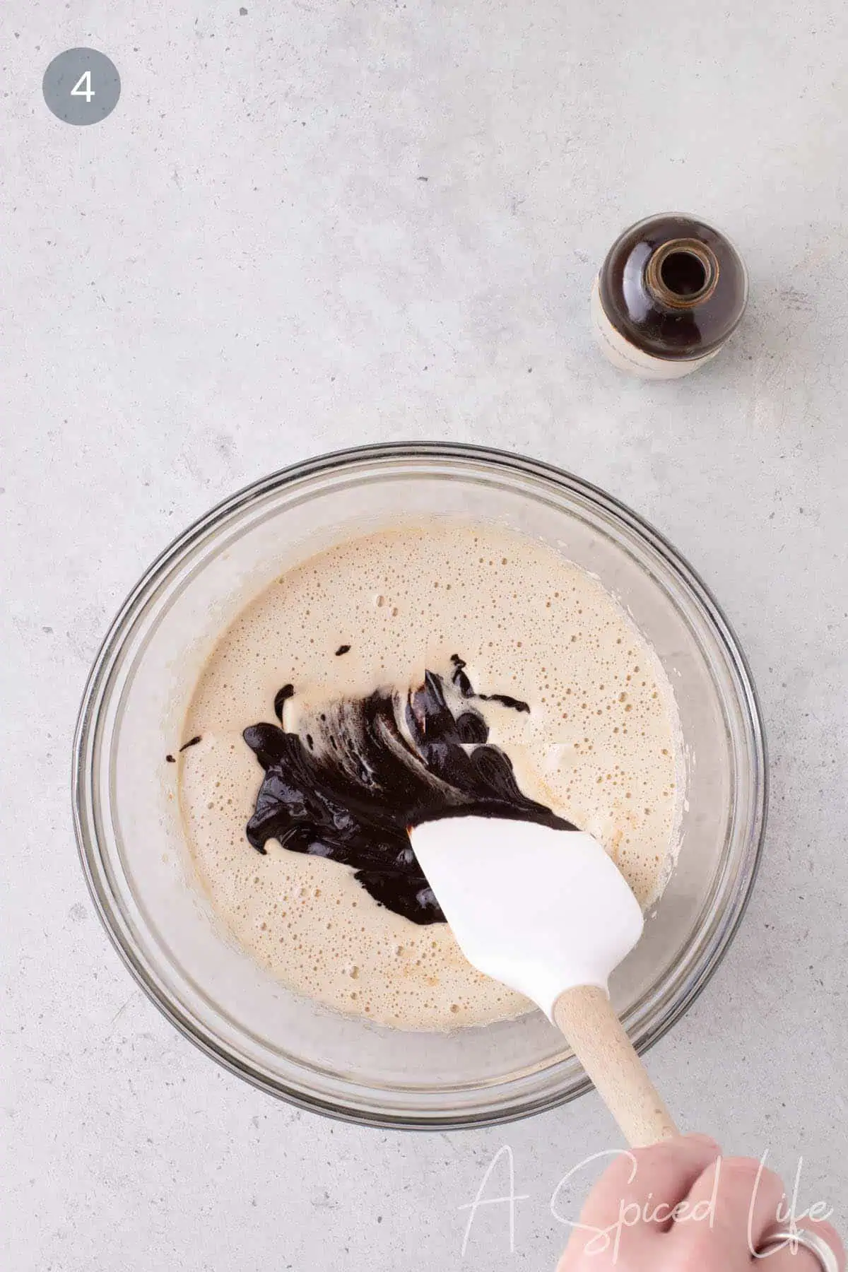Dark melted chocolate mixture being folded into a pale egg and sugar mixture in a glass bowl, with vanilla extract nearby.