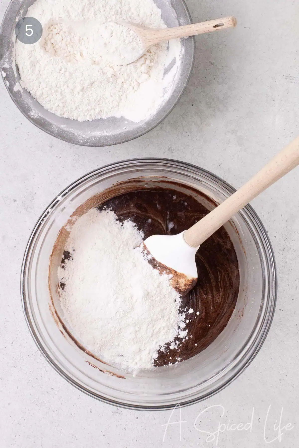 Flour mixture being folded into dark chocolate cookie batter with a spatula in a glass bowl.