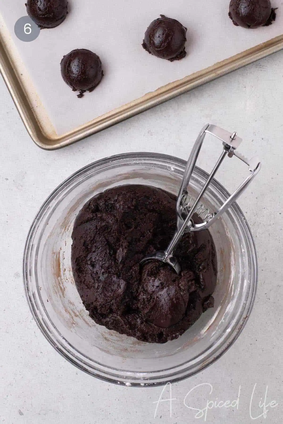 Chocolate cookie dough being portioned with a cookie scoop, with scooped dough on a parchment-lined baking sheet in the background.