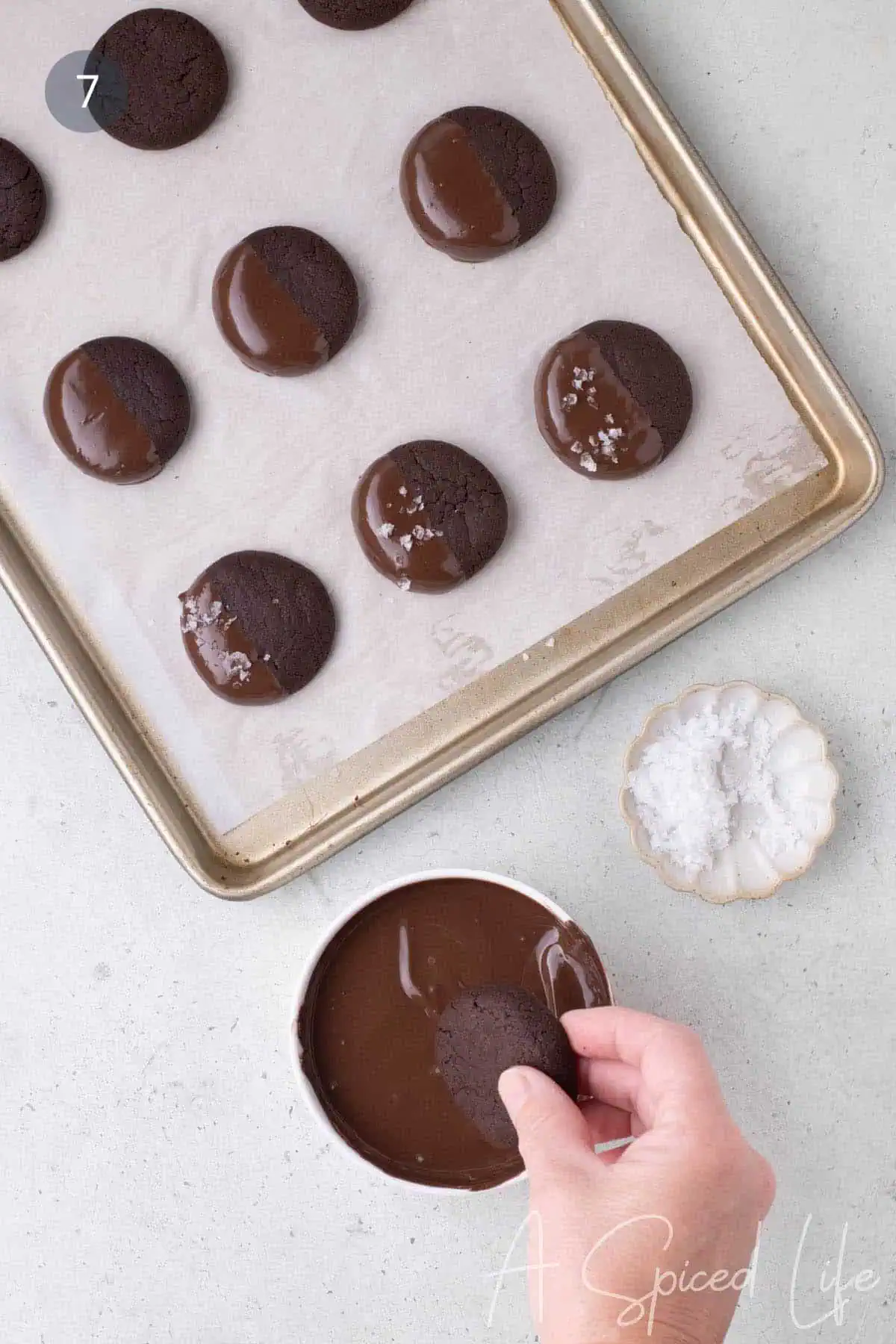 Chocolate cookie being dipped halfway into melted chocolate on a parchment-lined baking sheet.