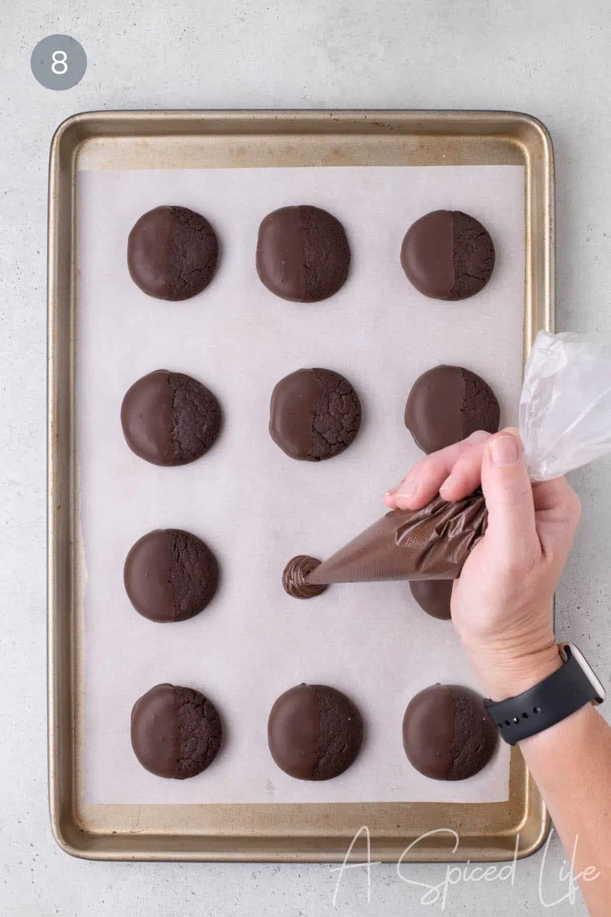 Melted chocolate being piped onto parchment beside half-dipped chocolate cookies.