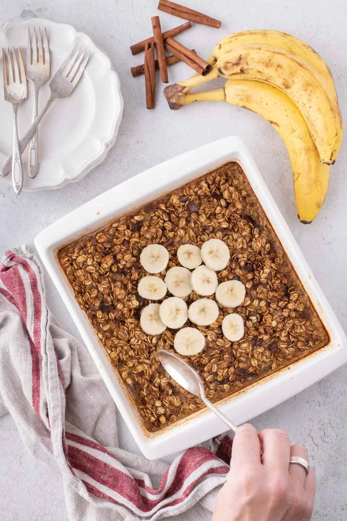 Baked banana bread oatmeal with dates and banana slices in a white baking dish, with a hand holding a serving spoon.