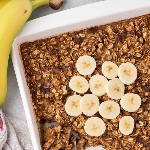 Banana-topped baked oatmeal in a white dish, with a portion scooped from one corner and a serving spoon resting inside.