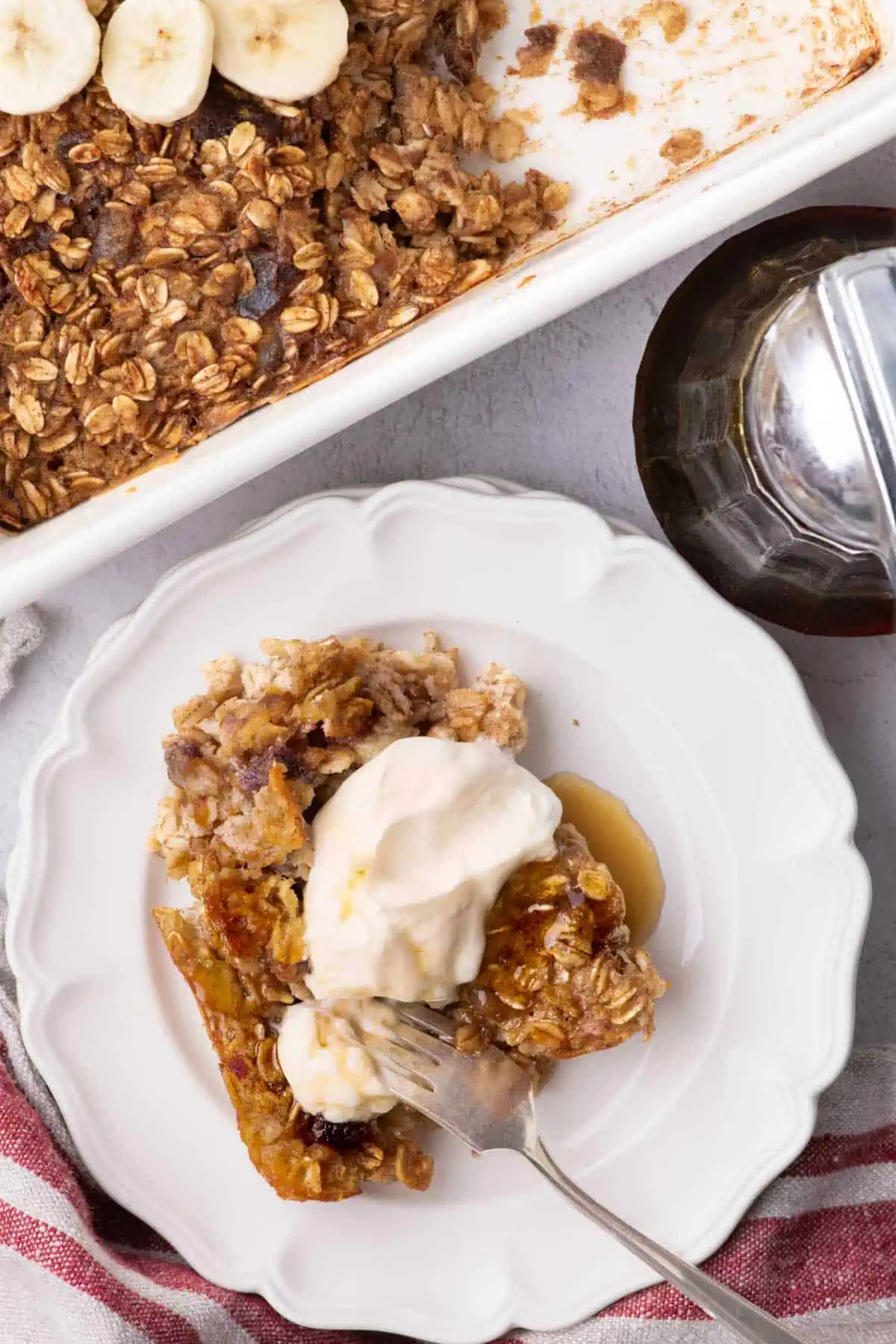 Overhead view of baked oatmeal with yogurt and maple syrup on a white plate beside the baking dish and syrup dispenser.