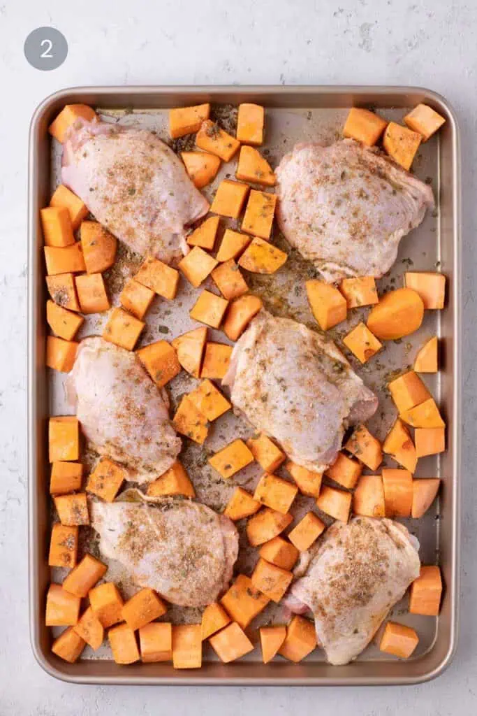 Bone-in chicken thighs and cubed sweet potatoes seasoned on a rimmed baking sheet before roasting.