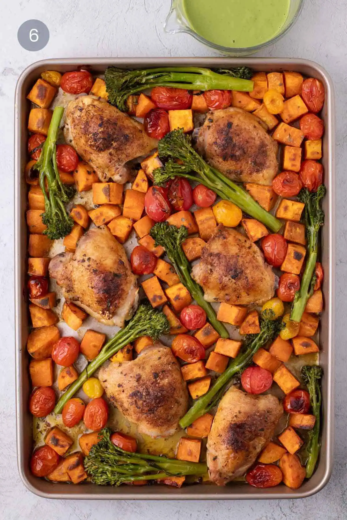 Roasted chicken thighs with caramelized sweet potatoes, broccolini, and cherry tomatoes on a sheet pan with a cup of cilantro cashew pesto.
