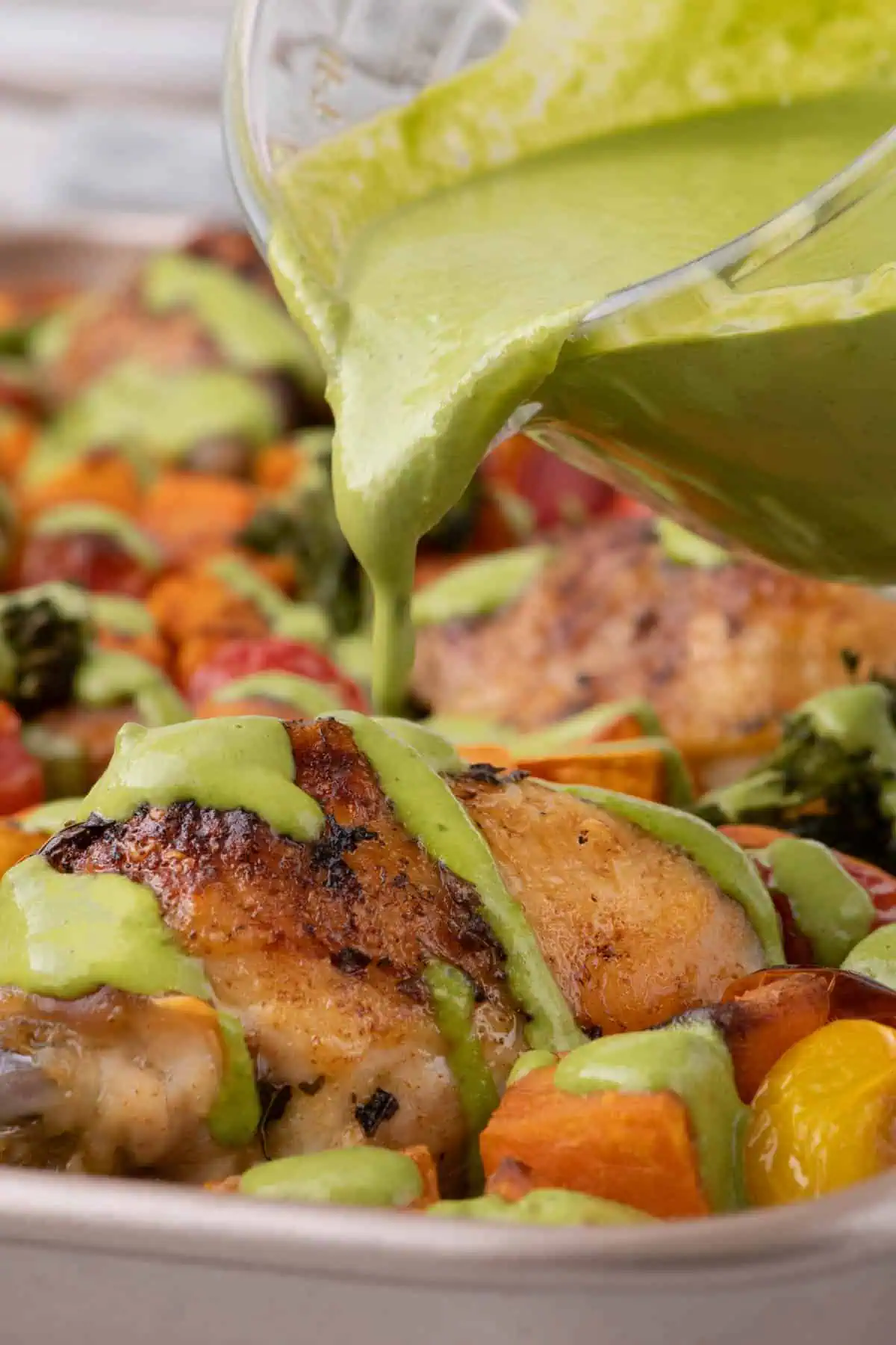 Cilantro cashew pesto being poured over crispy chicken thighs, sweet potatoes, broccolini, and roasted tomatoes on a sheet pan.
