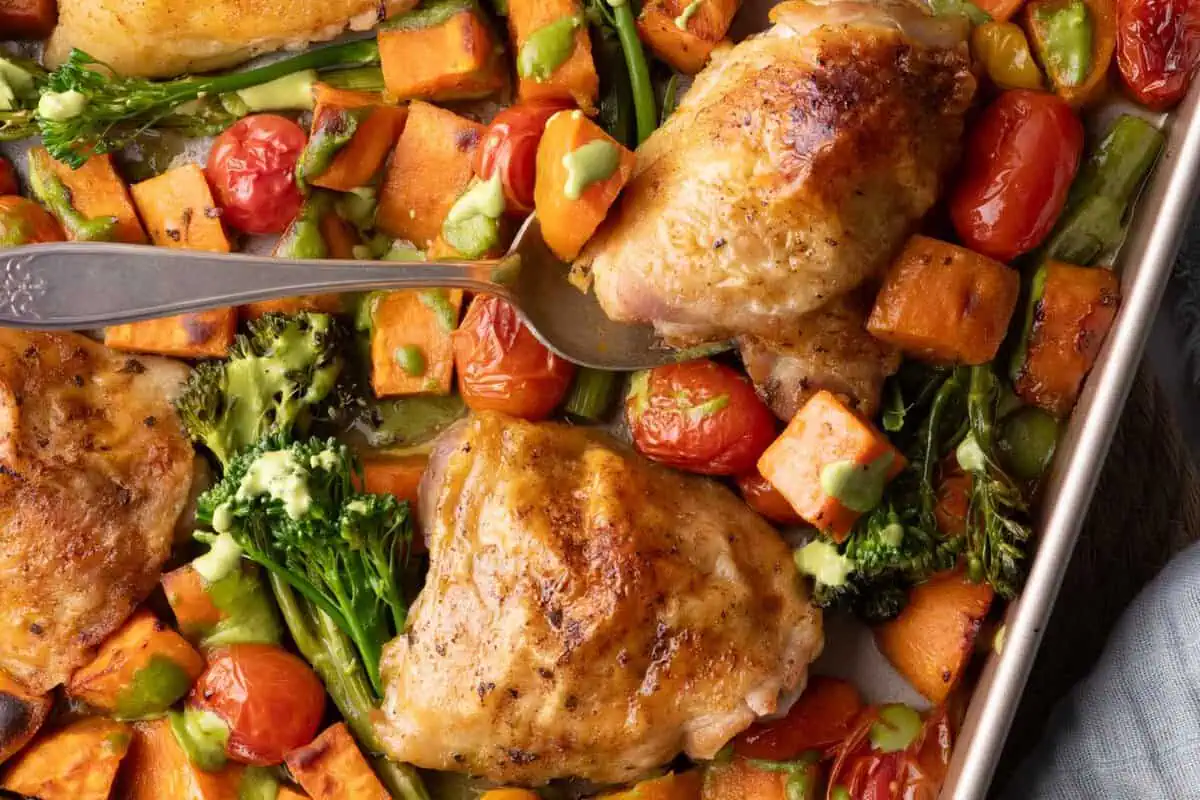 Crispy chicken thighs with roasted sweet potatoes, broccolini, and cherry tomatoes drizzled with cilantro cashew pesto on a sheet pan
