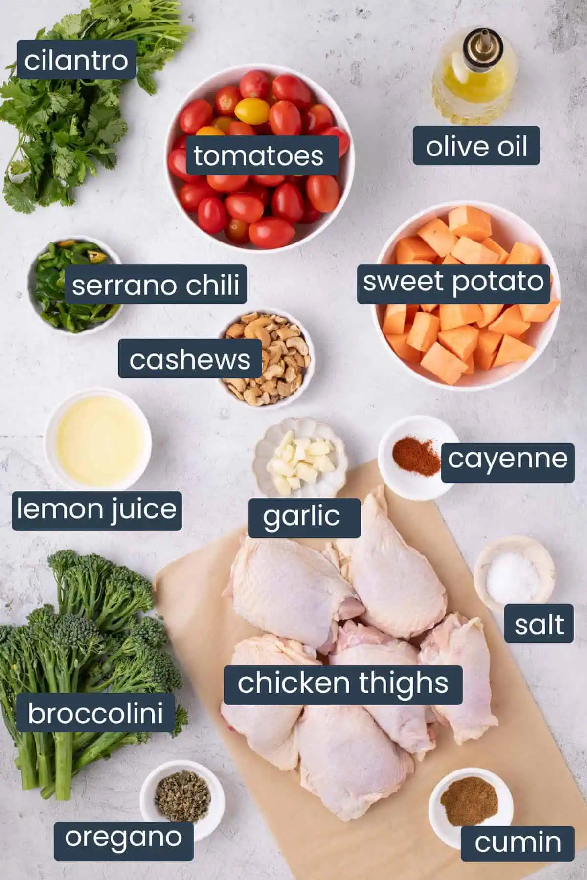Ingredients for chicken and vegetable sheet pan with cilantro cashew pesto, including chicken thighs, sweet potatoes, broccolini, tomatoes, cilantro, serrano chiles, cashews, garlic, lemon juice, olive oil, cumin, oregano, cayenne, and salt.